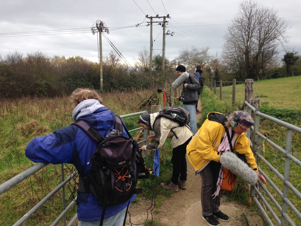 Five people next to metal barriers by a green field, making recordings with fluffy mics.